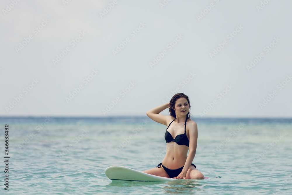 Fototapeta premium Beautiful surfing girl in bikini and with wet hairs sitting on a front of big longboard surf board and waiting for the wave in open ocean water. Modern active sport lifestyle and summer vacation.