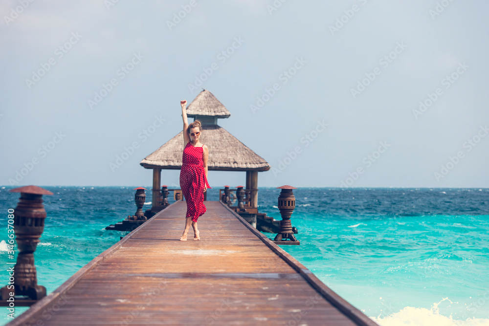 Joyful young happy girl in red jumpsuit standing on a wooden pier near water bungalows and enjoying ocean, summer breeze and sound of the waves during vacation. Happy holiday travel concept.