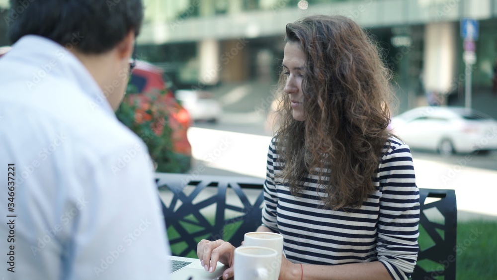Young woman in striped shirt and her handsome colleague in white shirt working together in summer park using laptop and clipboard. The man is drinking coffee Young woman in striped shirt and her handsome colleague in white shirt working together in summer park using laptop and clipboard. The man is drinking coffee