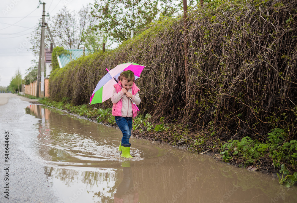 A beautiful little girl is walking down the street with a colorful umbrella and green rubber boots in the puddles. The girl walks down the street and looks at the puddles. A beautiful little girl is walking down the street with a colorful umbrella and green rubber boots in the puddles. The girl walks down the street and looks at the puddles.