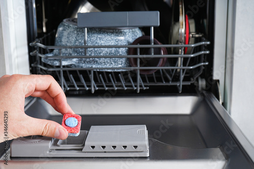 A man hand holding dishwasher detergent tablet. Putting tab into dishwasher, close up.  Dishwasher machine full loaded.