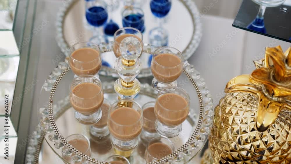 On a beautiful mirror tray on a buffet table are alcoholic, milk and coffee drinks of blue color in small glasses, next to them is a decorative glass in the form of a golden pineapple. On a beautiful mirror tray on a buffet table are alcoholic, milk and coffee drinks of blue color in small glasses, next to them is a decorative glass in the form of a golden pineapple.