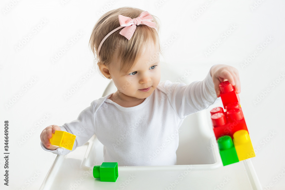 Nursery kid plays with educational toys in classroom sitting at the table in baby chair. Cute little girl playing colorful construction blocks. White background. Nursery kid plays with educational toys in classroom sitting at the table in baby chair. Cute little girl playing colorful construction blocks. White background.