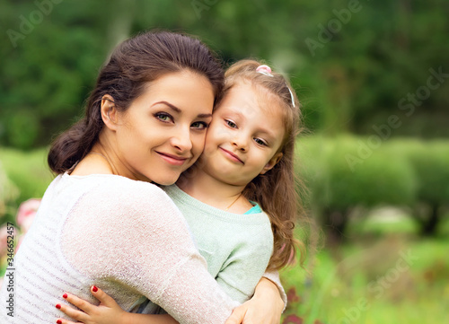 Beautiful mother hugging and kissing her cute small daughter on summer green grass background. Closeup