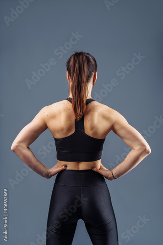 Pumped up strong fitness woman showing back and biceps muscles strength. Fit girl fitness model isolated on gray background. Hands up, hands on the waist line. Trainer wearing black sportwear
