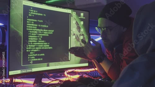 Lockdown of unrecognizable coder wearing hoody sitting at computer, coding and listening to bearded Caucasian man standing nearby leaning on desk giving comments concerning his code
