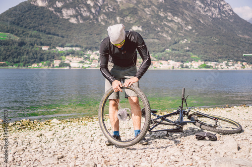Bike Repair. Man Repairing Mountain Bike. Cyclist man in trouble rear wheel wheel case of accident. Man Fixes Bike near lake in Italy background mountains. cyclist repairing bicycle wheel outdoors