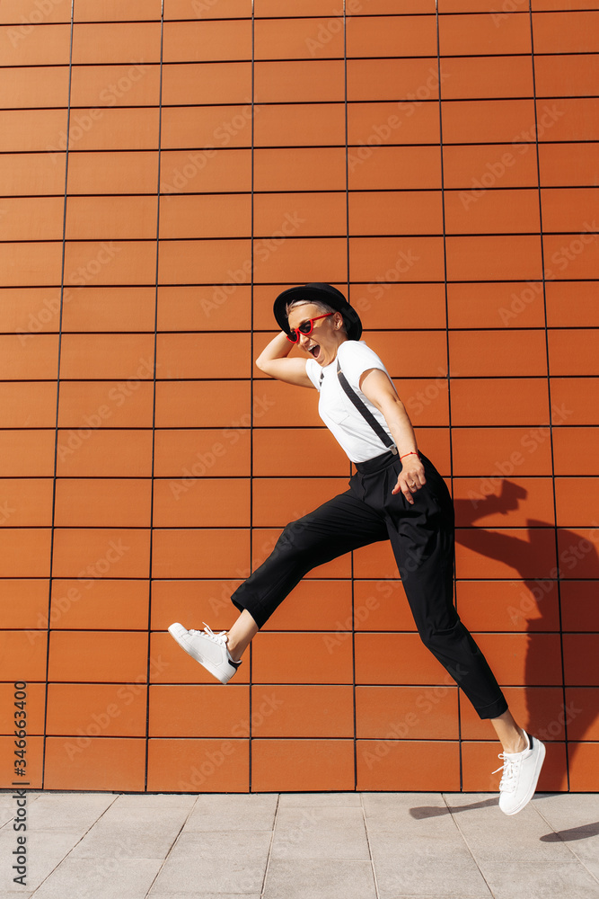Fototapeta premium Carefree hipster young woman wearing red sunglasses and black hat jumping near wall of modern building.