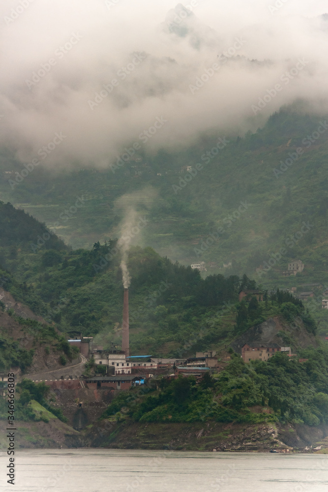 Xiangxicun, China - May 6, 2010: Xiling gorge on Yangtze River. Closeup of brown stone heavy industry plant with smokestack. Green mountains with descending cloudscape as backdrop.