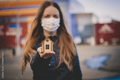 A girl in a medical mask holds a toy house in her hand. Stay home concept
