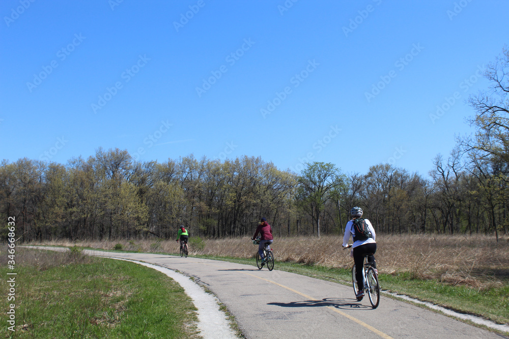 Obraz premium Three cyclists in spring on the North Branch Trail at Miami Woods in Morton Grove, Illinois practicing social distancing