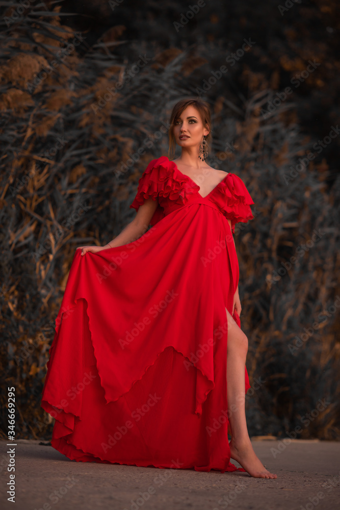lady stands in a luxurious red long dress barefoot on a green background of bushes, reeds