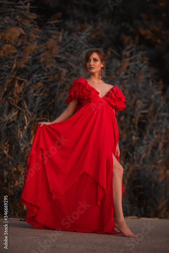 lady stands in a luxurious red long dress barefoot on a green background of bushes, reeds