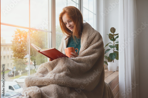 pretty thoughtful smiling young girl reading book and drinking morning coffee at home sitting next to the window wrapped in warm comfy blanket