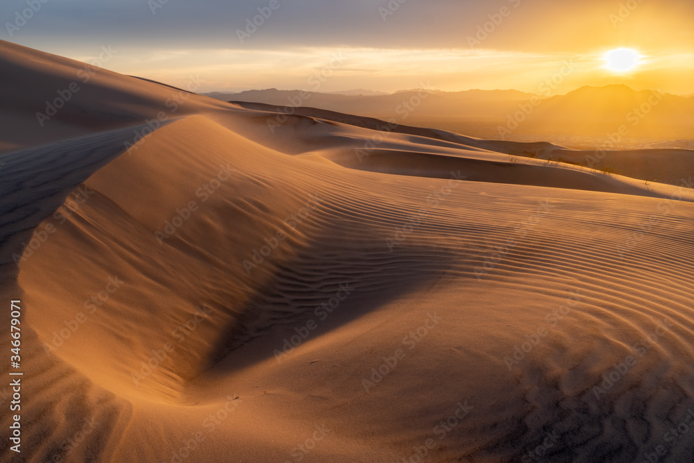 The desert sand is blowing with the strong evening wind creating waves and patterns in the dune as the sun is setting under cloudy sky over the mountains in the distance.