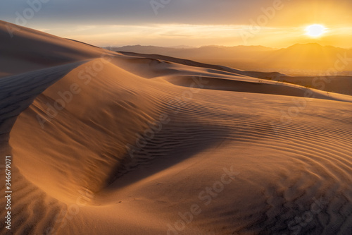 The desert sand is blowing with the strong evening wind creating waves and patterns in the dune as the sun is setting under cloudy sky over the mountains in the distance.