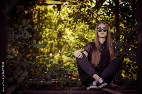 young girl in black clauses and glasses sit outside in the bushes