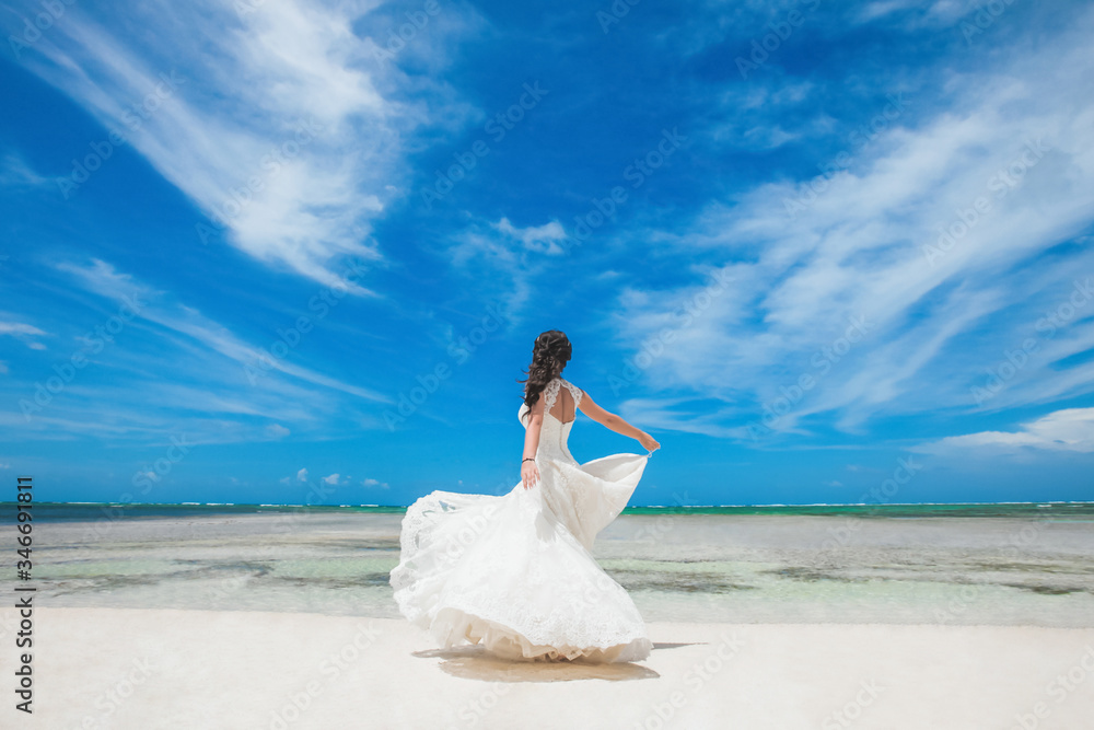 Naklejka premium Bride in a white wedding dress with a veil walking on the sandy caribbean beach at sunny day in Dominican republic