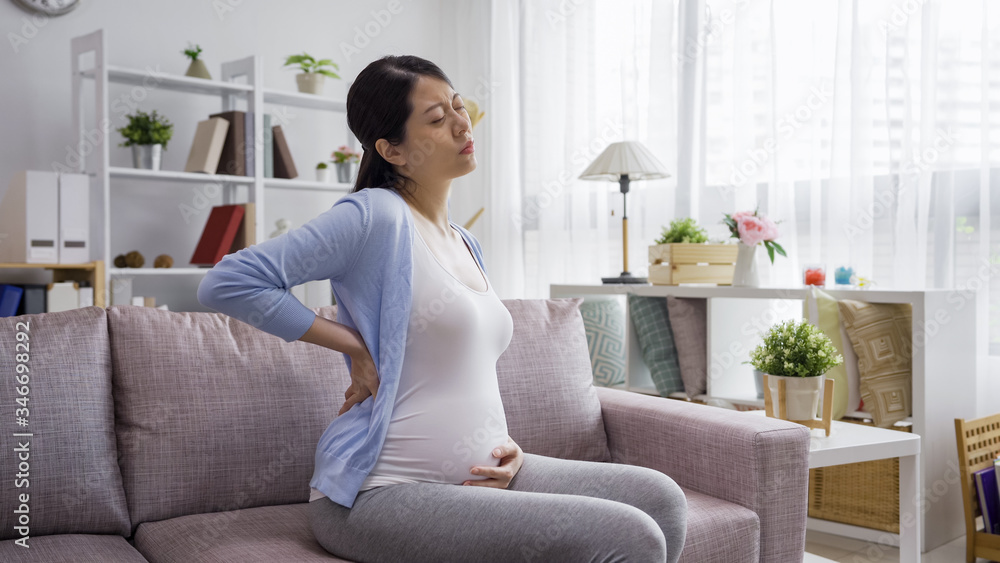 upset pregnant woman hand hold abdomen and touch waist on couch. frowning asian chinese lady future parenthood having back aches in the last trimester of pregnancy. Concept of pregnancy health care.