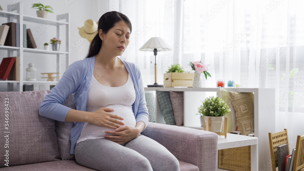 Young pregnant woman suffering from abdominal pain at home. future motherhood holding big belly and feels hurt while sitting in couch in bright living room in house. illness mom hands on tummy Young pregnant woman suffering from abdominal pain at home. future motherhood holding big belly and feels hurt while sitting in couch in bright living room in house. illness mom hands on tummy