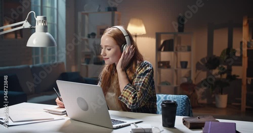 Positive redhead caucasian high school or university student listening to music in headphones and concetrating on work. Young freelancer enjoying remote job in laptop 4k footage photo