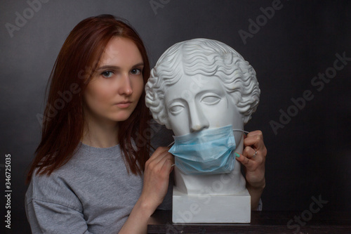 Red-haired young girl a black background a gray t-shirt and plaster bust in protective medical mask