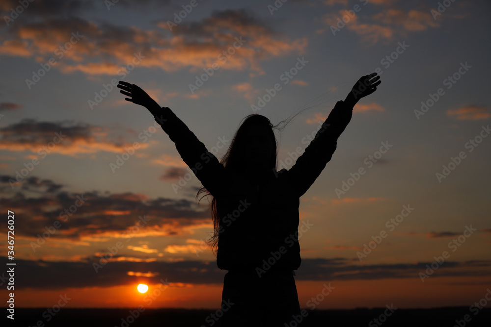 The young girl feels happy and raises her hand. Sunset time. Silhouette of a young girl with hands up at sunset.