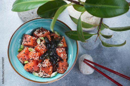 Poke bowl with salmon over light-blue stone background with green ficus leaves and pebbles, high angle view, horizontal shot