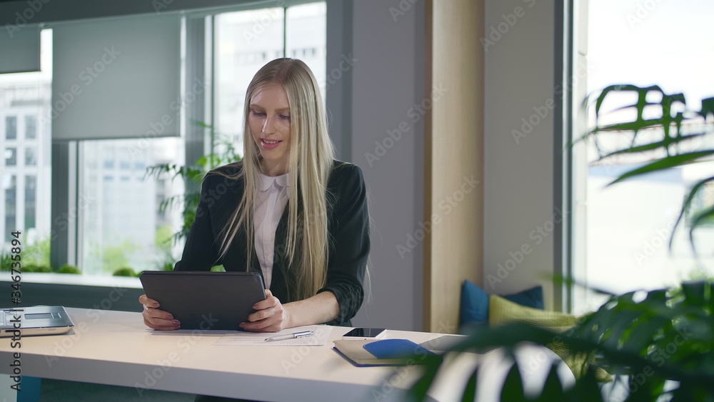 Elegant woman in suit sitting at table with laptop and surfing tablet in modern light office with big windows Elegant woman in suit sitting at table with laptop and surfing tablet in modern light office with big windows