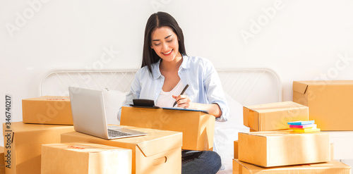 Young smiling beautiful owner asian woman freelancer sme business online shopping working on laptop computer with parcel box on bed at home - SME business online and delivery concept