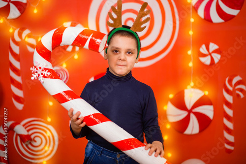 A child with reindeer horns on his head holds in his hands a large toy lollipop standing on a background of garlands, lollipops and sweets.