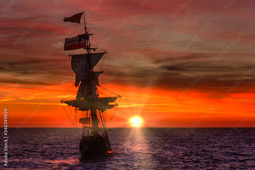 Fototapeta premium Antique wooden ship leaving the harbor under a bright red sunset spring time