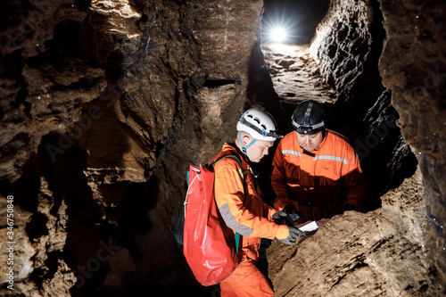 Two men, strong physique, explore the cave. Men dressed in special clothes to pass through the cave and stopped, looking at the map photo