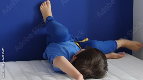 three-year-old child in blue pants and a T-shirt jumps on a white bed mattress against the wall in the apartment