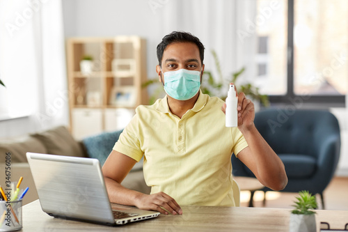 quarantine, remote job and pandemic concept - happy indian man wearing face protective medical mask for protection from virus disease with hand sanitizer and laptop computer working at home office