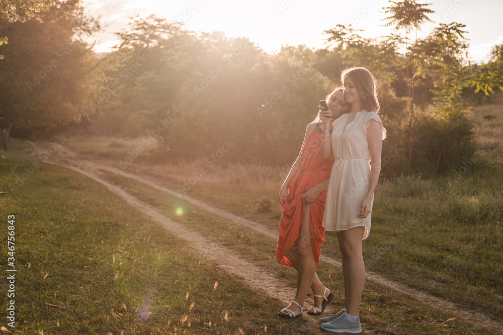 Two girlfriends walk together in the woods at sunset. It is beautiful summer weather. Both are dressed in dresses. Two girlfriends walk together in the woods at sunset. It is beautiful summer weather. Both are dressed in dresses.