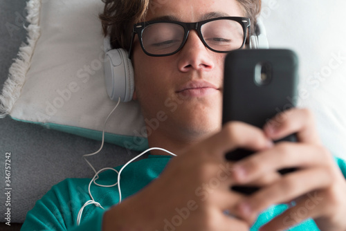 Young handsome caucasian teenager use cellular phone device and listen music with headphones lay down at home - download songs activity and modern connected life concept