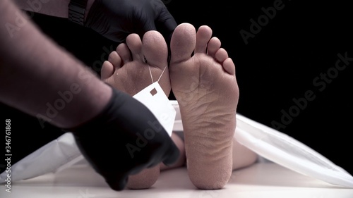 Pathologist in black gloves hangs tag with sign COVID-19 on foot of corpse. Finger move of zombie. legs of girl or guy who died from illness are in morgue under white blanket. Dry skin on the feet.