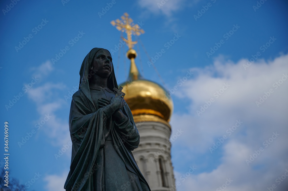 Fototapeta premium A statue of a praying nun against the background of an Orthodox church in Donskoy monastery in Moscow. A golden cross on a blue sky background.