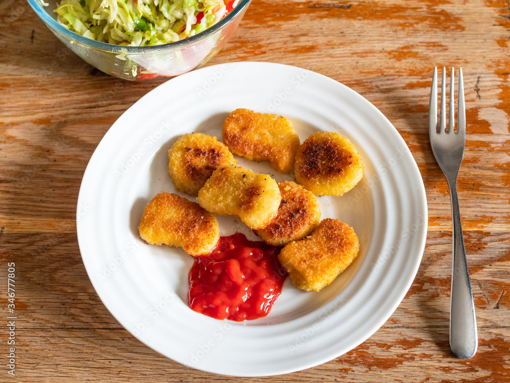 cooked Chicken nuggets from frozen product (deep-fried breaded chicken meat) with tomato ketchup on white plate and salad from fresh vegetables on old wooden table in home kitchen cooked Chicken nuggets from frozen product (deep-fried breaded chicken meat) with tomato ketchup on white plate and salad from fresh vegetables on old wooden table in home kitchen