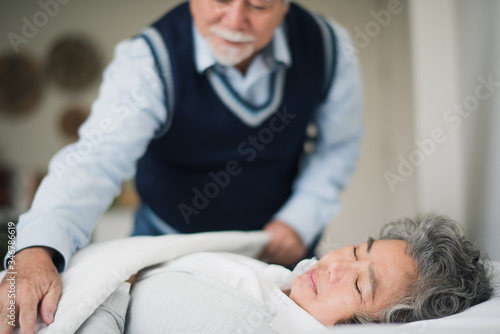 Senior Asian couple love lying on the bed. Retired man take care by blanket woman when sleeping on bed in bed room at home which smiling and felling happy. Old couple love concept.
