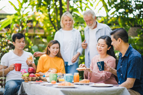 Happy big family have a lunch at outdoor in green garden. Lunch or tea time on picnic table in summer. Old woman make a bread with Fruit jam to give husband. Big family outdoor lunch concept.