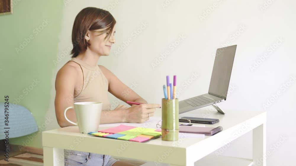 Young woman works, greets, communicates online via webcam, talks, laughing, talking about important things, learns an online conference. Home decor, sitting at table, short hair, wearing beige top. Young woman works, greets, communicates online via webcam, talks, laughing, talking about important things, learns an online conference. Home decor, sitting at table, short hair, wearing beige top.