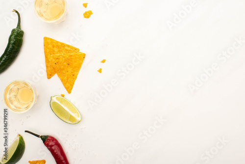 top view of golden tequila with lime, chili peppers, nachos on white marble surface