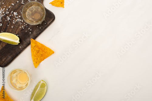 top view of golden tequila with lime, salt and nachos near wooden cutting board on white marble surface