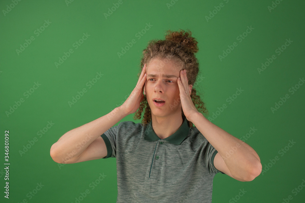 The first signs of coronavirus. Headache in a man. The guy holds his head grimacing in pain. Curly guy on a green background looking at the camera. The first signs of coronavirus. Headache in a man. The guy holds his head grimacing in pain. Curly guy on a green background looking at the camera.