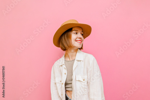 Happy tourist girl in hat and light clothing isolated on pink background, looks away at copy space and smiles. Lady in summer clothes stands on pink wall background. Vacation concept.