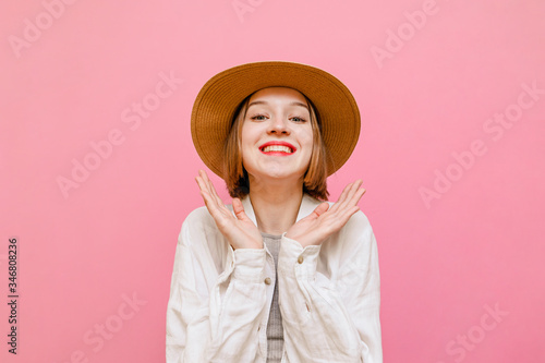 Portrait of joyful lady with happy face and clenched fists cheers, looks at camera and smiles. Cute girl in hat and light clothing rejoices on pastel pink background. Isolated.