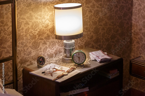 comodino illuminato da un alampada con occhiali da vista cruciverba orologio e sveglia, su parete con carta d sparati vintage photo