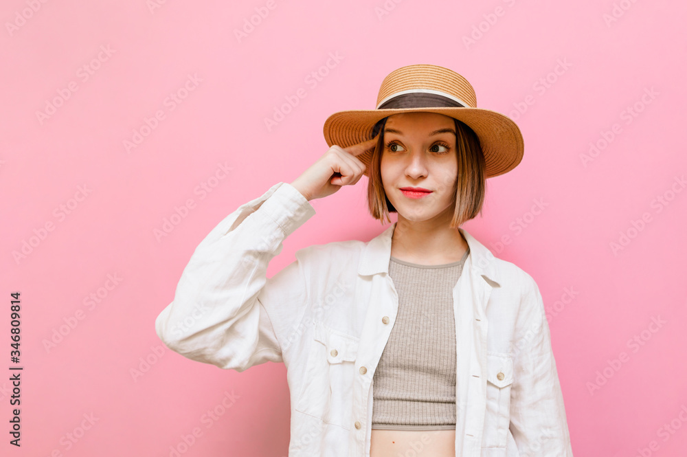 Emotional girl in hat and light clothing stands on pink background with finger attached to temple, looks away at copy space. Surprised lady looking away at shows gesture 